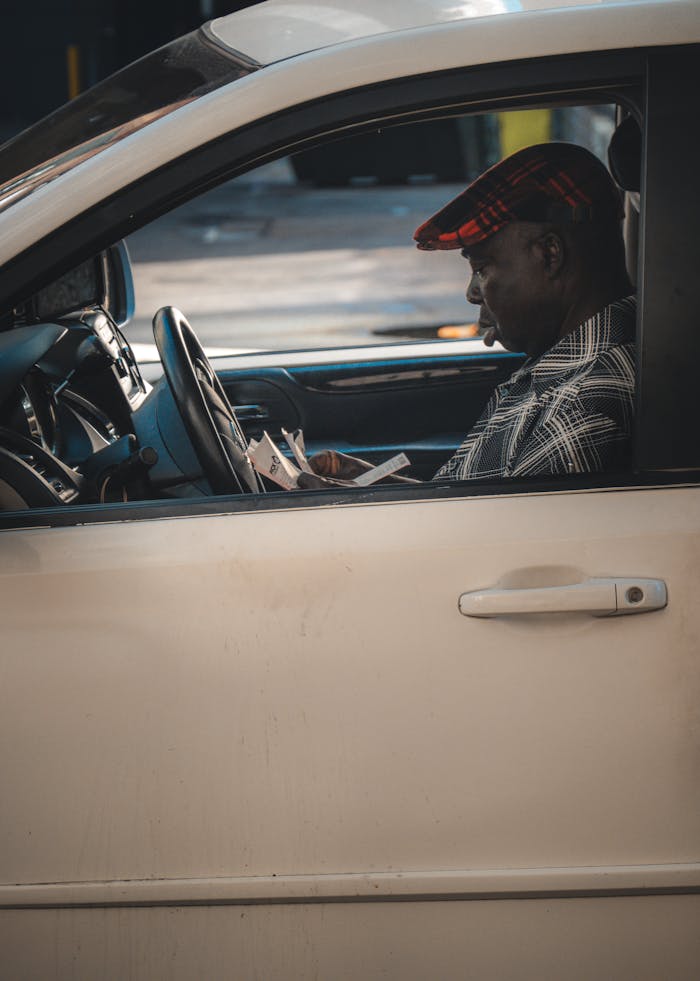An adult man reads a newspaper inside a parked car during daytime.