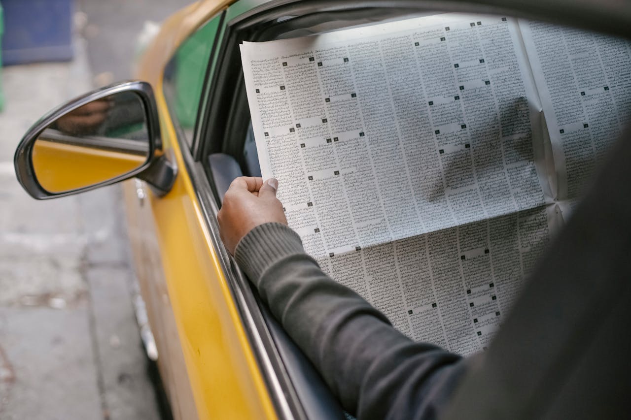 A person reads a newspaper through a car window, capturing a moment of leisure and information.